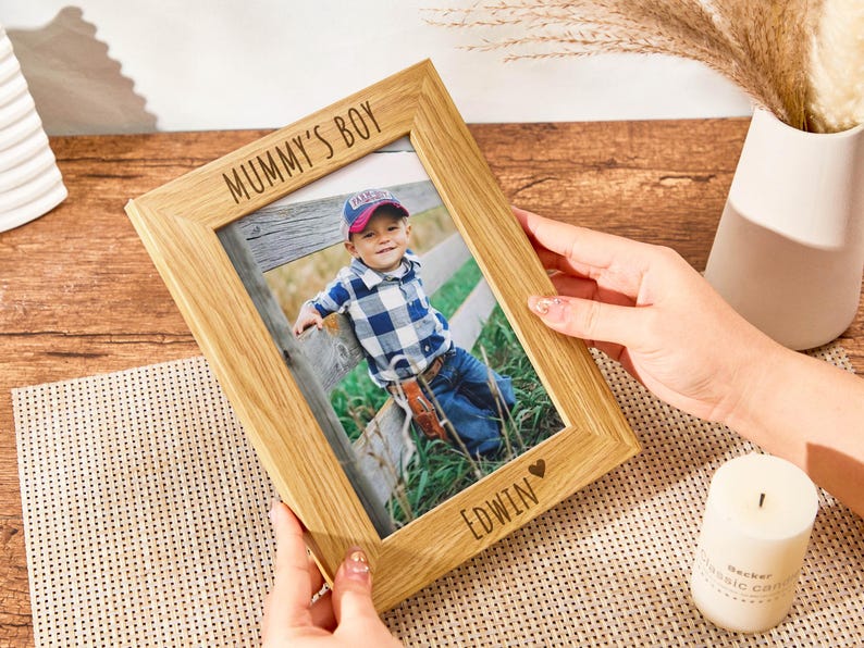 May include: A wooden photo frame with a light brown finish. The frame has a white inscription that reads "Mummy's Boy" and a smaller inscription that reads "EDWIN" with a heart symbol. The frame contains a photo of a young boy wearing a plaid shirt and jeans.
