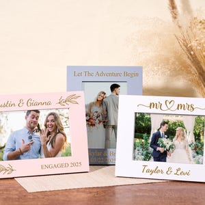 May include: Three framed photos with wedding and engagement themes. One pink frame says "Justin & Gianna ENGAGED 2025". A blue frame says "Let The Adventure Begin". A white frame says "mr & mrs Taylor & Levi".