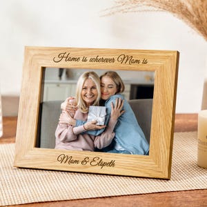 May include: A wooden picture frame with a photo of two people hugging. The frame is engraved with the text "Home is wherever Mom is" and "Mom & Elspeth".