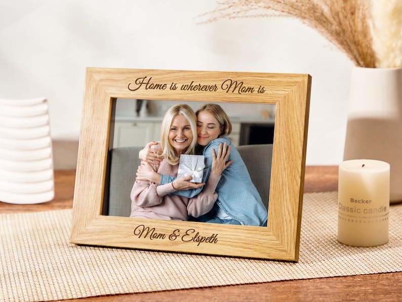 May include: A wooden picture frame with a photo of two people hugging. The frame is engraved with the text "Home is wherever Mom is" and "Mom & Elspeth".