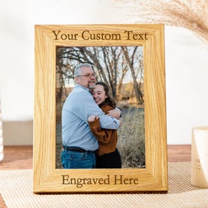 May include: A wooden picture frame with a photo of two people hugging. The frame has a space for engraving with the text "Your Custom Text" and "Engraved Here".