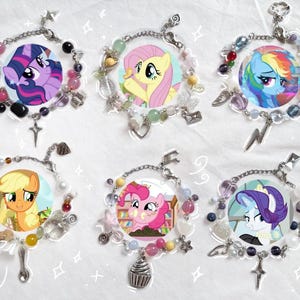 May include: Six colourful charm bracelets featuring My Little Pony characters. Each bracelet has a different pony, including Twilight Sparkle, Pinkie Pie, Rainbow Dash, Fluttershy, Applejack, and Rarity. The bracelets are made with silver chains, beads, and charms.
