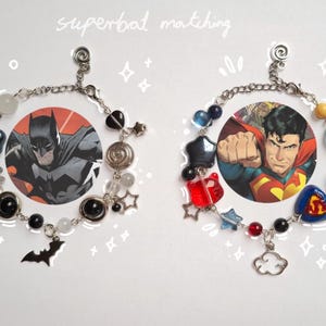 Könnte beinhalten: Zwei passende Armbänder mit Comicfiguren. Eines hat ein Batman-Bild, schwarze Sternperlen und einen Fledermaus-Anhänger. Das andere hat ein Superman-Bild, rote und blaue Perlen und einen Wolken-Anhänger. Der Text "superbat matching" steht darüber.