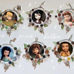 Può includere: Sei ciondoli circolari con personaggi delle fate, ciascuno incorniciato con metallo argentato, perline e accenti di foglie. I ciondoli presentano immagini di fate. La parola "pixie hollow" è scritta in corsivo.