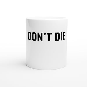 Könnte beinhalten: Weißer Keramikbecher mit dem Aufdruck "DON'T DIE" in fetten schwarzen Buchstaben. Der zylindrische Becher steht auf einer weißen Oberfläche. Ein Becher in Standardgröße für Heißgetränke.