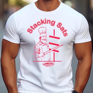 Puede incluir: Camiseta blanca con un gráfico rojo de un chef sosteniendo un cuchillo y las palabras "Stacking Sats". El chef lleva un gorro blanco alto y una chaqueta de chef. El diseño está centrado en la parte delantera.