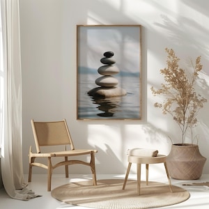 May include: A framed print of a stack of smooth, gray and white stones balanced on top of each other in a body of water. The stones are reflected in the water. The print is hanging on a white wall in a room with a wooden chair and a small stool.
