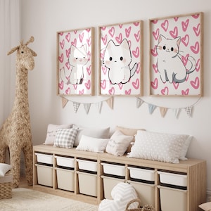 Puede incluir: Tres impresiones enmarcadas con fondos rosas y gatos blancos con corazones. Las impresiones están colgadas en una pared blanca sobre un banco de madera con contenedores de almacenamiento.