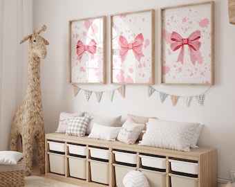May include: Three framed art prints featuring watercolor pink bows on a white background with pink splatter details. A woven giraffe and storage bench with pillows and bins complete the nursery decor.