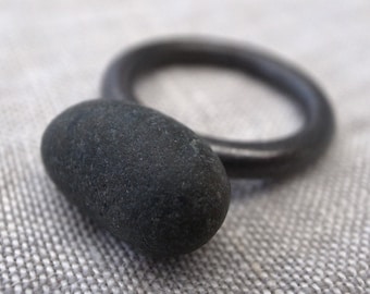 Puede incluir: Una piedra ovalada de color gris oscuro junto a un anillo oscuro. La piedra tiene una superficie texturizada y es lisa al tacto. El anillo es circular y parece ser de metal. Los objetos están sobre un fondo neutro y texturizado.