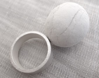 Puede incluir: Un anillo de plata y una esfera blanca con una superficie texturizada. Ambos objetos están sobre un fondo de tela gris claro.