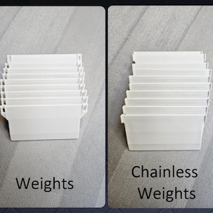 May include: A set of white vertical blind weights and a set of white chainless vertical blind weights. Each set consists of multiple rectangular plastic components with small attachment hooks at the top, arranged in a neat, stacked vertical row.