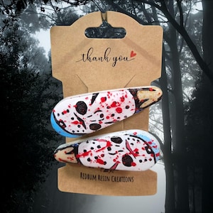 May include: Two hair clips featuring a white hockey mask design with black eye holes and red blood splatter accents. The clips are attached to a brown cardboard display card with the text thank you and Redrum Resin Creations printed on it.