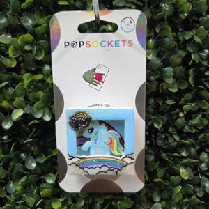 May include: A square PopSockets phone grip featuring a My Little Pony character with a rainbow mane and tail. The design includes a light blue frame, a glittery finish, and a rainbow graphic at the base. It is mounted on a white retail display card.