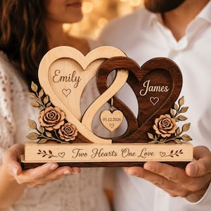 Puede incluir: Una pieza decorativa de madera con dos corazones entrelazados, uno claro y otro oscuro, grabados con los nombres Emily y James. Un pequeño corazón cuelga en el centro con la fecha 05.12.2026. La base está grabada con la frase Two Hearts One Love.