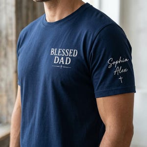 Może przedstawiać: Granatowa koszulka z białym napisem "BLESSED DAD" na piersi, z krzyżem poniżej. Na rękawie widnieją imiona "Sophia Alex" i krzyż. Codzienna, swobodna koszulka.