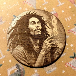 Könnte beinhalten: Ein brauner Holz-Untersetzer mit einem lasergravierten Bild von Bob Marley, der eine Zigarette raucht. Das Bild ist im Vintage-Stil gehalten und hat einen warmen Braunton.
