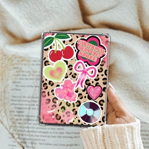 Puede incluir: Una funda para Kindle de estampado de leopardo rosa y negro con varias pegatinas, incluyendo cerezas, corazones, un lazo, un ángel y un disco. El texto en una pegatina dice "Emotional Support Kindle".