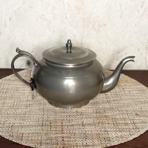 Op de afbeelding: Een zilverkleurige theepot met een gebogen tuit en handvat. De theepot heeft een deksel met een kleine knop bovenop. Hij staat op een ronde, geweven placemat. De achtergrond is een getextureerde, gebroken witte muur.