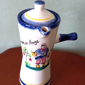 Puede incluir: Jarra de cerámica blanca con asa y boquilla azules, con tapa con ribete azul y banda amarilla. La jarra está decorada con una colorida ilustración de una persona y texto.