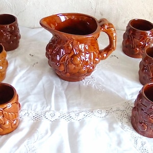 Peut inclure: Une cruche en céramique marron et six tasses assorties. La cruche a une anse et un bec, et les tasses ont des anses. Toutes les pièces sont décorées d'un motif de feuilles et de baies en relief.