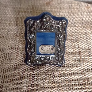 Pode incluir: Uma pequena moldura prateada ornamentada com um fundo de veludo azul. A moldura apresenta um design detalhado de querubins e volutas. O texto "GENUINE SILVER PLATED MADE IN ENGLAND" é visível.