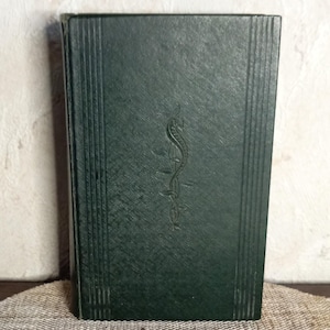 Op de afbeelding: Een donkergroen boek met een getextureerde kaft en verticale lijnen op de rug. Een medisch symbool is in reliëf op de voorkant aangebracht. Het boek staat rechtop op een neutrale ondergrond.