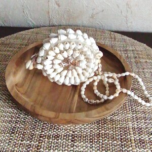 Op de afbeelding: Een decoratief boeket gemaakt van kleine witte schelpen, rustend op een houten dienblad. Een schelpenketting is naast het boeket gedrapeerd. De lade staat op een geweven mat, waardoor een natuurlijk, kustthema ontstaat.
