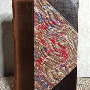 Puede incluir: Libro antiguo con lomo y esquinas de cuero marrón. La cubierta presenta un diseño veteado en rojo, azul y gris. Los bordes del libro están desgastados, lo que sugiere edad. Una pieza clásica para coleccionistas.