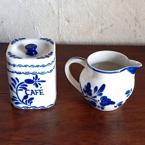 Peut inclure: Service à café en céramique blanche avec des motifs floraux bleus. L'ensemble comprend un pot carré avec un couvercle et le mot "CAFE" et une petite cruche à crème. L'ensemble est sur une surface en bois.