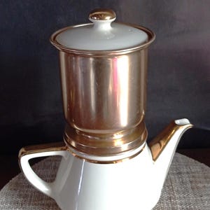 Op de afbeelding: Een vintage koffiezetapparaat met een witte keramische basis en een goudkleurig metalen bovendeel. De koffiepot heeft een wit deksel met een gouden knop en gouden rand. De tuit en het handvat zijn ook goudkleurig.