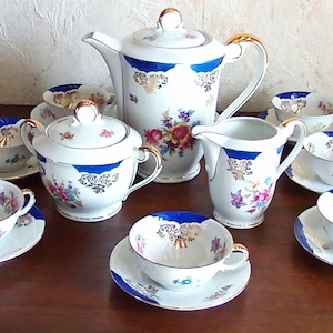 Peut inclure: Service à thé en porcelaine vintage avec fond blanc, bordures bleues et motifs floraux. L'ensemble comprend une théière, un pot à lait, un sucrier et plusieurs tasses avec soucoupes. Des accents dorés ornent les poignées et les bords.