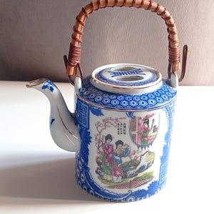 Peut inclure: Théière décorative en céramique avec un motif floral bleu et blanc et une anse tressée. La théière présente une illustration détaillée de personnages dans une scène de jardin. Le bec et le bord sont rehaussés d'une garniture dorée.