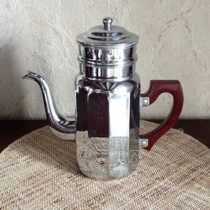 Op de afbeelding: Een vintage, gepolijste chromen koffiepot met een donkerbruine houten handgreep. De pot heeft een rechthoekige body met verticale groeven en een deksel met een kleine knop. Hij staat op een geweven, ronde placemat.