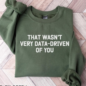 May include: A military green crewneck sweatshirt featuring the white text "THAT WASN'T VERY DATA-DRIVEN OF YOU" printed across the center of the chest. The garment has long sleeves with ribbed cuffs and a ribbed neckline, showcasing a solid color design.
