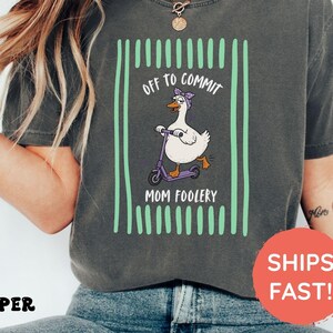 Op de afbeelding: Een antracietgrijs T-shirt met korte mouwen en een afbeelding van een witte gans met een paarse bandana op een paarse step. De tekst OFF TO COMMIT MOM FOOLERY wordt omlijst door verticale lichtgroene strepen op een donkere achtergrond.