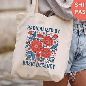 Op de afbeelding: Een crèmekleurige canvas tas met een grafische print van rode bloemen en blauwe bladeren. De tekst "RADICALIZED BY BASIC DECENCY" is in blauwe hoofdletters boven en onder het bloemmotief op de voorzijde van de tas gedrukt.