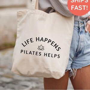 May include: A cream-colored canvas tote bag featuring the black printed text LIFE HAPPENS PILATES HELPS with a small lotus flower icon centered between the two lines of text. The bag has two shoulder straps and a simple rectangular shape.