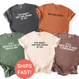 May include: Five t-shirts in a variety of colours, including grey, orange, green, cream, and brown. Each shirt has a different text-based design in white, with phrases such as "Talk Data to Me" and "It's a Beautiful Day to Analyse Data."