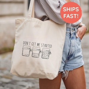 May include: Cream-colored tote bag with the phrase "DON'T GET ME STARTED" printed in black. Below the text are three jars with lids and tags. The bag is carried over the shoulder.