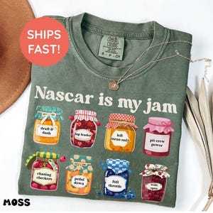 Peut inclure: T-shirt vert mousse avec l'inscription "Nascar is my jam" et des illustrations de pots de confiture avec des étiquettes sur le thème de la course. Le t-shirt a un col rond et des manches courtes. Un collier doré est drapé sur le t-shirt.