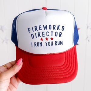 May include: A red, white, and blue trucker hat with the text "FIREWORKS DIRECTOR I RUN, YOU RUN." The hat has a red brim, a white front panel, and a blue mesh back. A red button is at the top.