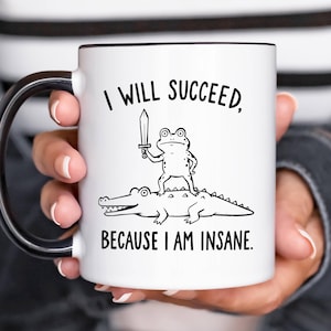 May include: A white ceramic mug with a black handle and rim, featuring a black line drawing of a frog holding a sword while standing on an alligator. The text above reads I WILL SUCCEED, and the text below reads BECAUSE I AM INSANE.