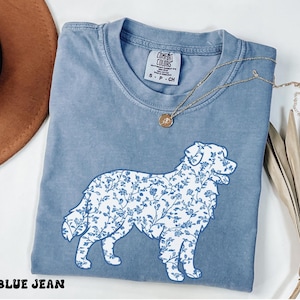 May include: A blue jean colored t-shirt featuring a white graphic of a dog filled with a blue floral pattern. The shirt has a crew neckline and a Comfort Colors brand label on the inside collar. A gold necklace with a small round pendant rests on top.