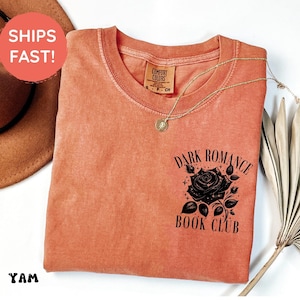 May include: A terracotta-colored short-sleeve t-shirt featuring a black graphic on the chest. The graphic depicts a rose surrounded by stars and the text "DARK ROMANCE BOOK CLUB". The shirt has a crew neckline and a Comfort Colors brand label.