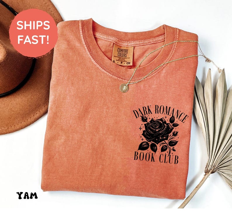 May include: A terracotta-colored short-sleeve t-shirt featuring a black graphic on the chest. The graphic depicts a rose surrounded by stars and the text "DARK ROMANCE BOOK CLUB". The shirt has a crew neckline and a Comfort Colors brand label.