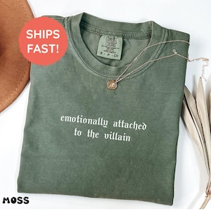 May include: A moss green t-shirt with the text "emotionally attached to the villain" in white. A gold-colored necklace with a pendant is draped on the shirt. The shirt has a Comfort Colors tag. A brown hat is visible in the background.
