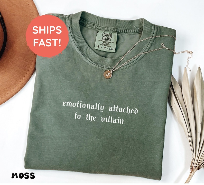 May include: A moss green t-shirt with the text "emotionally attached to the villain" in white. A gold-colored necklace with a pendant is draped on the shirt. The shirt has a Comfort Colors tag. A brown hat is visible in the background.