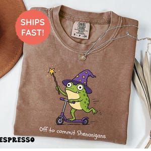 Può includere: Una t-shirt color espresso con la grafica di una rana verde che indossa un cappello da mago viola con stelle gialle, su un monopattino viola mentre tiene in mano una bacchetta magica. Il testo sotto recita, Off to commit Shenanigans.