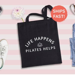 May include: A black canvas tote bag featuring two shoulder straps and white printed text that reads LIFE HAPPENS PILATES HELPS, centered above a small lotus flower icon. The bag is displayed against a light pink and white striped background.